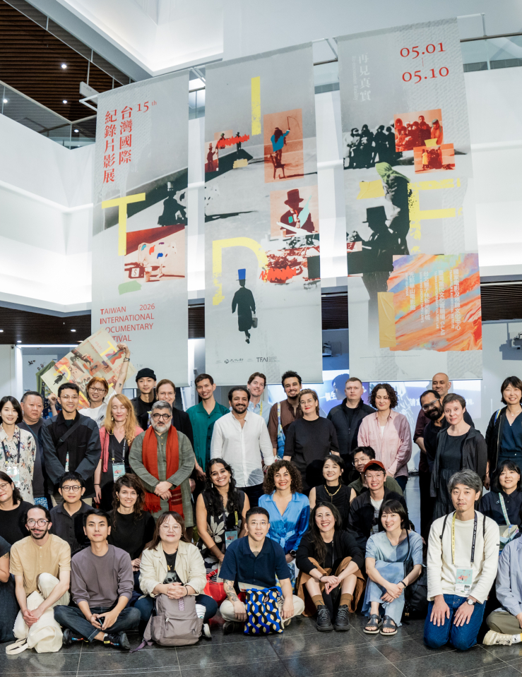 50 people of varying ages and races stand and kneel in three rows in a large, open exhibition space. Three large banners behind the group read "Taiwan International Documentary Festival 2026"