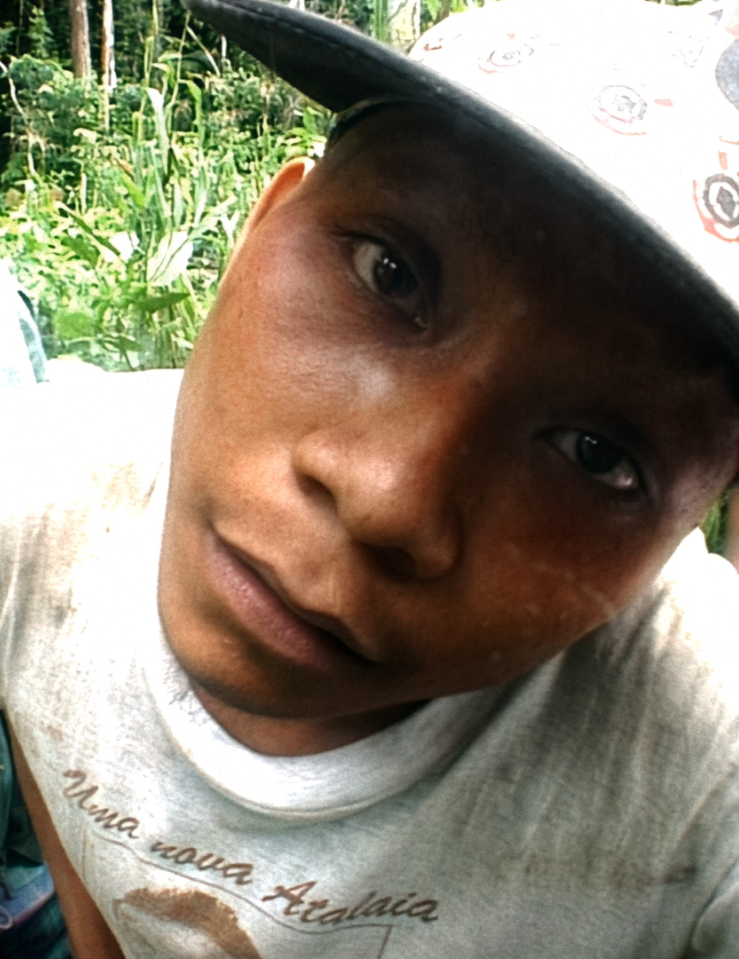 A man in a cap stares directly into the camera. The angle is distorted, as if shot with a fish eye lens. Another man in the background also looks into the camera. The two are in a densely vegetated area