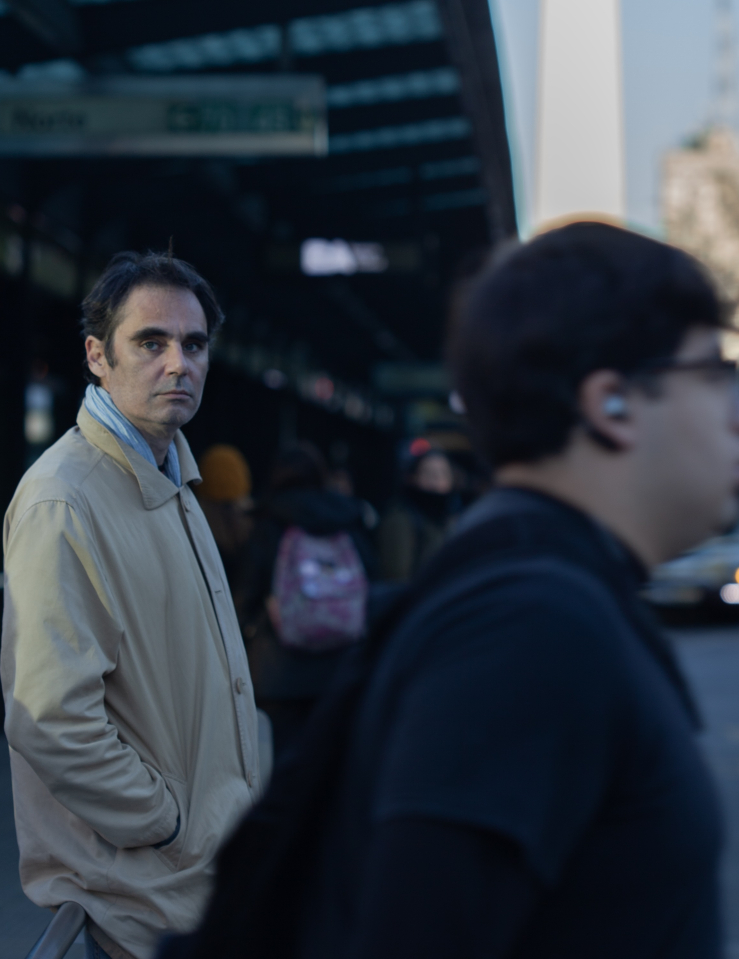 Middle-aged white man in a khaki rain coat over a light blue shirt stands in a somewhat crowded street looking straight at the camera