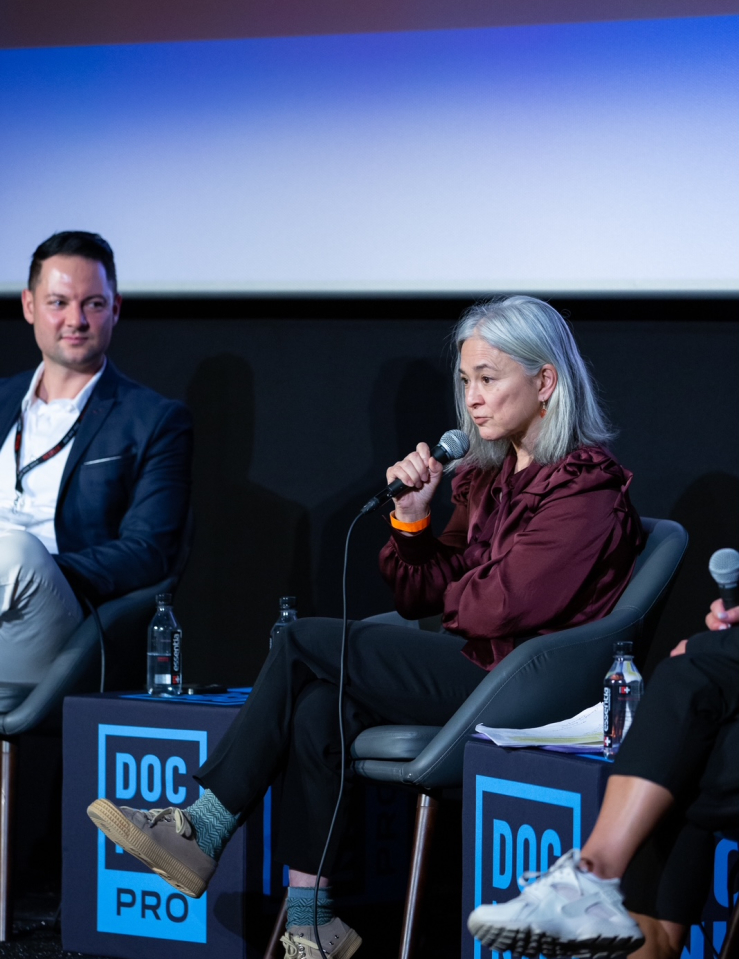 A woman in a maroon button-down shirt speaks into a microphone. She sits on a chair infront of a large screen. A man in a white shirt and blue blazer sits to her right. Both face forward. Tables on either side and in-between the two read "DOC NYC PRO"