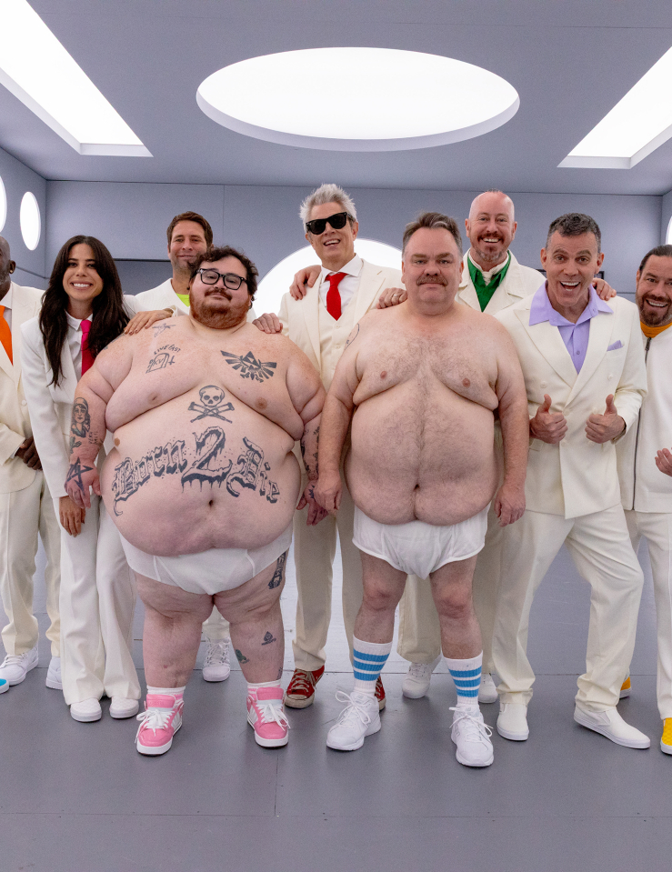 Two large men wear nothing but sneakers and diapers. One is decorated in tattoos, including a skull and crossbones and text which reads "BORN 2 DIE." Nine men and one woman stand behind the two in matching white suits. The ensemble appears in a sterile, grey room with futuristic, circular white lights