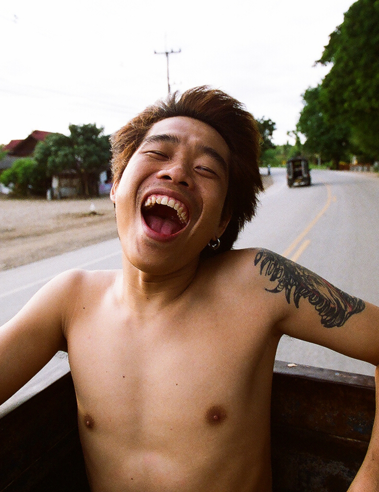 Young shirtless Asian man with dyed light red hair smiling broadly while on the bed of a moving truck on the road