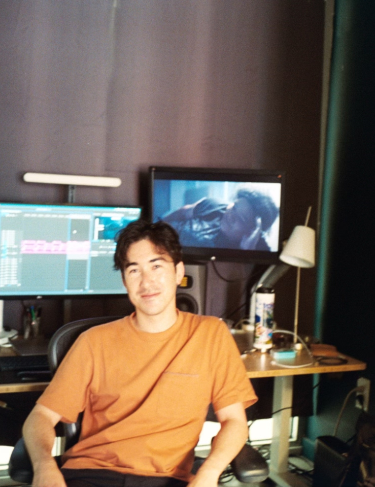 Young man in orange shirt sits in front of editing workstation with two large monitors