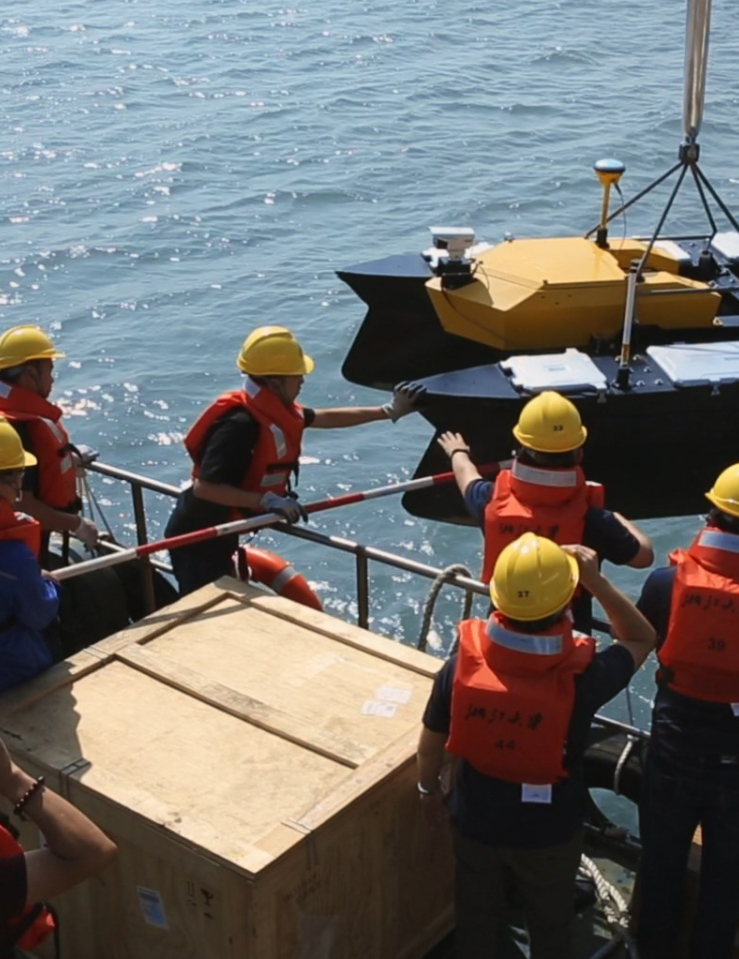 Eight men in orange vests and yellow hard hats on ship raise two containers out of the ocean