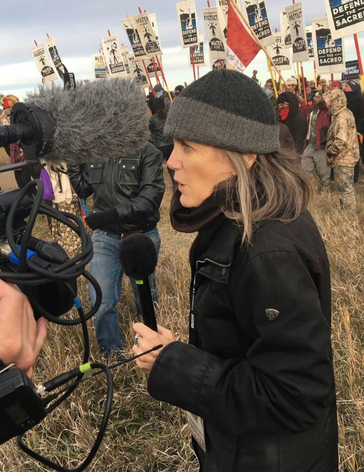 A middle-aged white woman in a black jacket and a grey wool hat stands in front of a protest holding a mic speaking into a camera