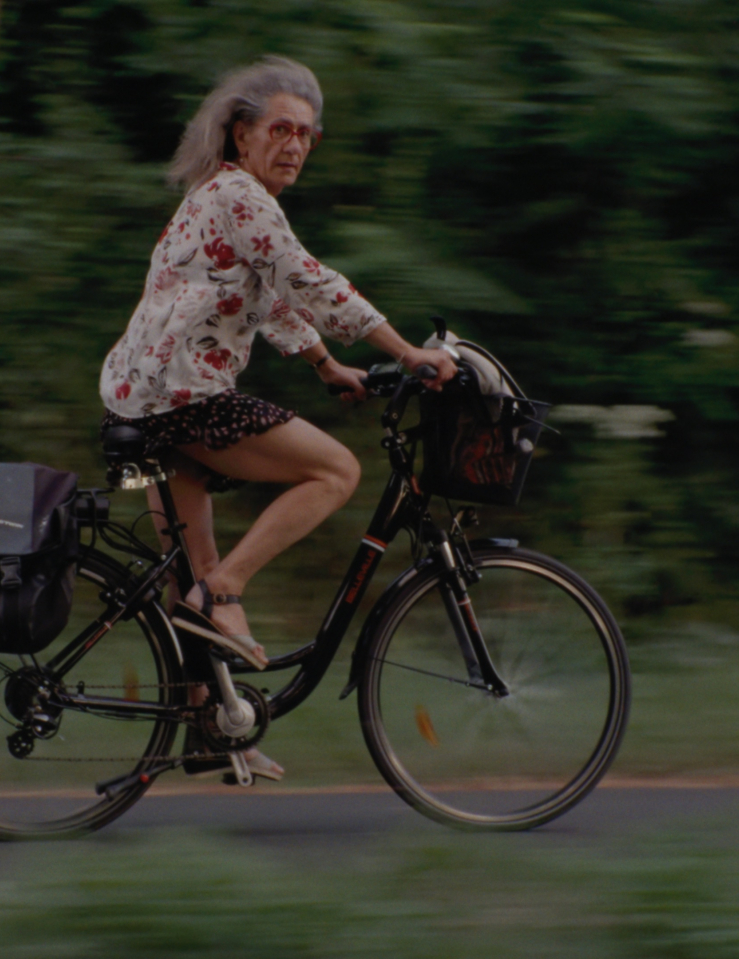 A woman with long grey hair, red glasses, a floral top, a short black skirt and sandals rides a black bike with a black bike bag along a forested path while looking to her right at the camera