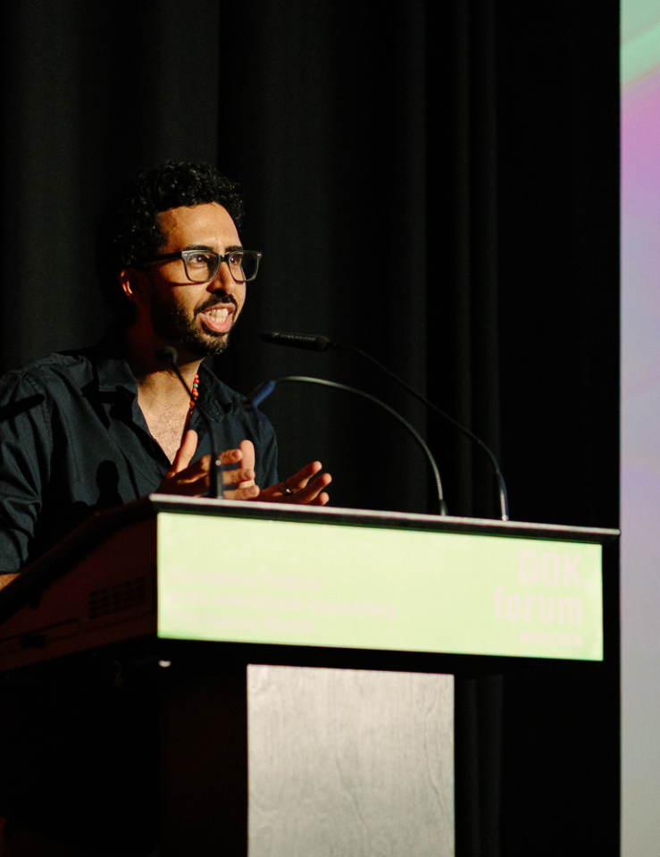 A man in a black shirt with black glasses and short dark hair and beard delivers a speech at a podium. Behind him is a black curtain and the lefthand side of a pink and green digital display showing text which cannot be read
