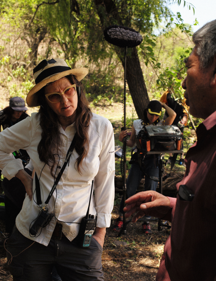 A woman (Lucrecia Martel) in a white button down shirt with glasses and a straw hat, wearing a walkie talkie and lanyard, speaks to a man in a red shirt. Behind the two are a film crew of three, one man holding a makeshift fill, one woman holding a boom pole and microphone and sound monitoring system, and one person mostly obscured by Martel