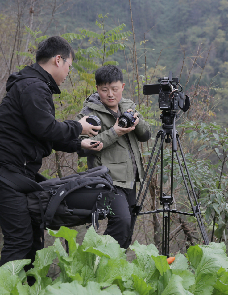 A man in a green jacket trades lenses with a man in all black. They stand behind a camera on a tripod amid forest