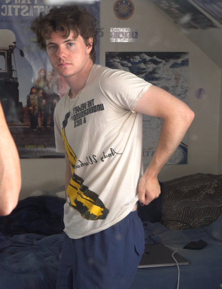 A young white man with short curly dark hair in an Andy Warhol white tee stares at a mirror in his bedroom as a white woman whose face is obscured by a camera takes a photo of them both