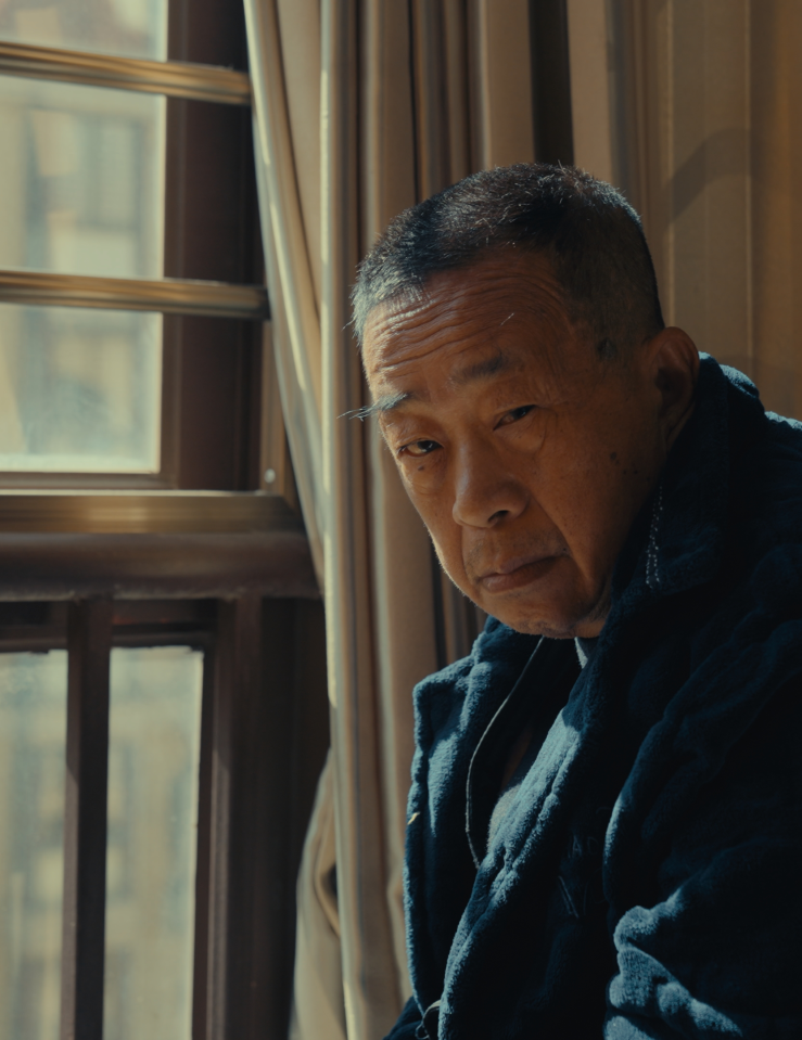 An aging Asian man with short-cropped hair in a bulky winter jacket glares at the camera while hunched over