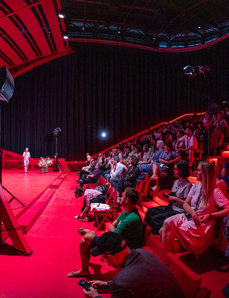 A large crowd in a theater lit in red looks at a big screen which displays a map of Czechia over a purple background