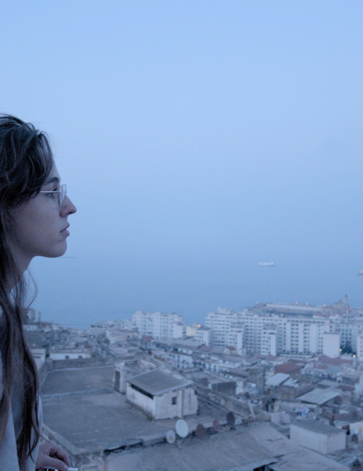 A woman in glasses with long brown hair looks over a city on the ocean at dusk