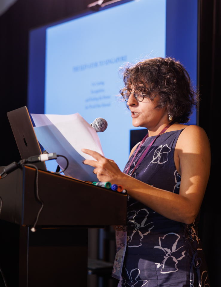 A woman (Sarvnik Kaur) with short, curly hair in a floral dress and glasses stands at a podium, speaking into a microphone. Behind her, out of focus, are a sign language interpreter and a large screen displaying text on a white background.