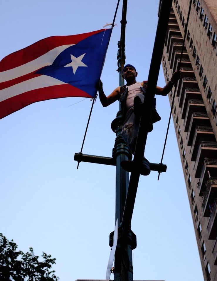 A man stands on a telephone pole with a large Puerto Rican flag flying next to him. Shot from beneath the pole, and the background is filled with the sky and one side of an apartment building