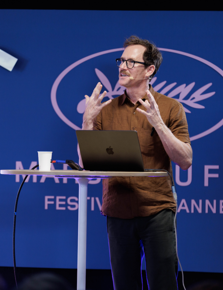 White middle-aged man with brown hair, glasses and a neat mustache wears a brown shirt against a blue backdrop at Cannes's Marche du Film standing in front of a laptop mid-performance