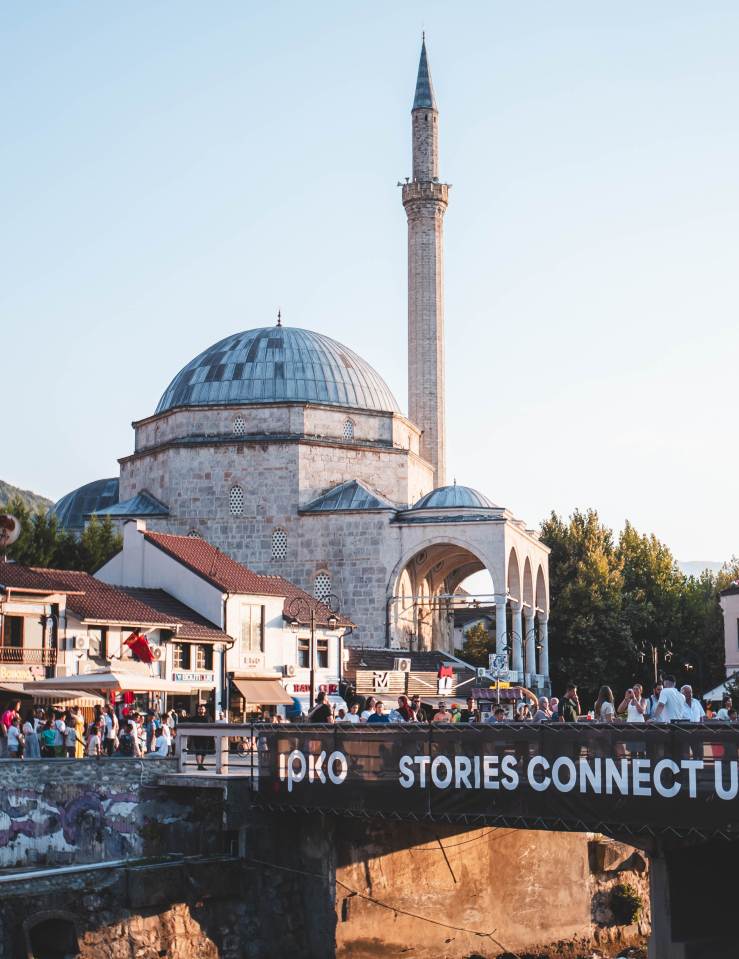 A place of worship in the city of Prizren, Kosovo, with a bridge in front of it from which a banner hangs reading "STORIES CONNECT US DOKUFEST." People flood the streets