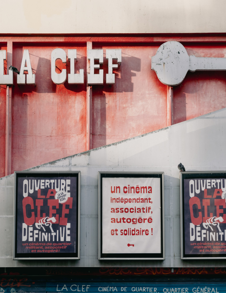 The front of a building, half red and half grey. At the top are the words "LA CLEF" and the shape of a key in grey. Three large posters are displayed on the building, reading "Ouverture La Clef Dēfinitive" and and "Un cinēma indēpendant, associatif, autogērē et solidaire!"