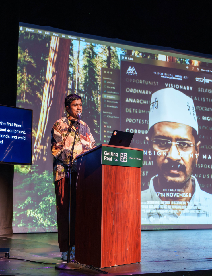 A man (Vinay Shukla) in a colorfully patterned shirt stands behind a podium and speaks into a microphone on stage. Behind him, a screen displays a poster of "An Insignificant Man" and an ASL interpreter, and a smaller screen provides captions of his words