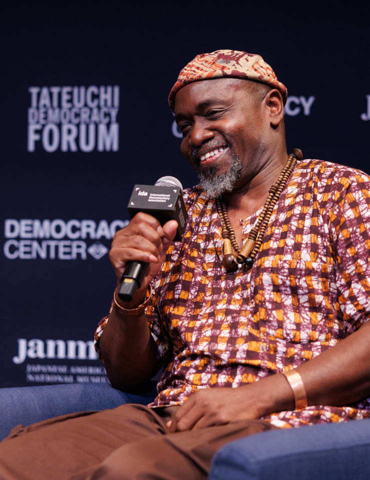 A smiling man in a shirt and hat composed of the same orange, brown, and white patterned fabric, wearing beads around his neck and a metal bracelet, holds a microphone labelled "ida international documentary association" and sits in a blue armchair. Behind him is a black background with white text reading "TATEUCHI DEMOCRACY FORUM", "janm", and "DEMOCRACY CENTER"