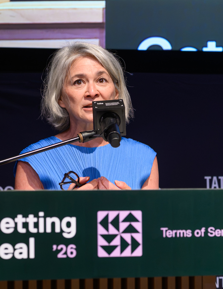 A woman in a blue dress (Chang) speaks into a microphone behind a podium which displays a green sign reading "Getting Real" in white text and "'26 Terms of Service" in pink text
