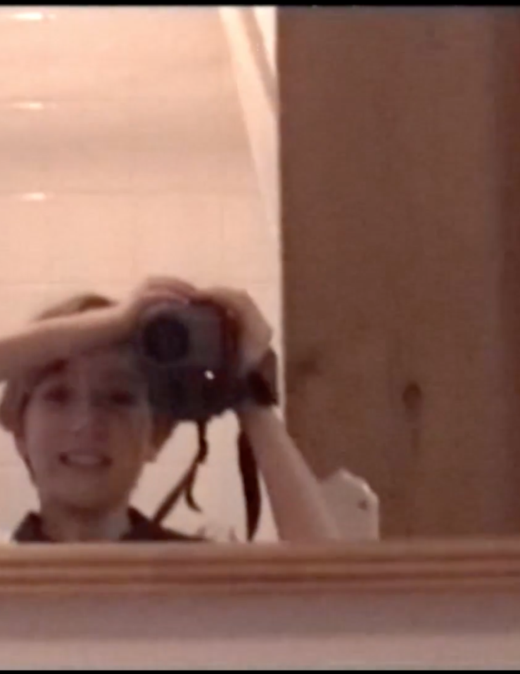 Pink tinted image of a smiling little girl with short hair filming herself in a mirror