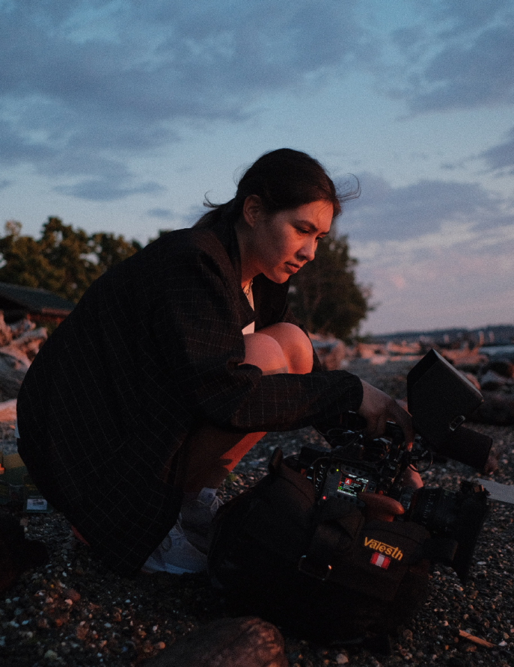 A young woman squats on a beach holding a film camera at dusk