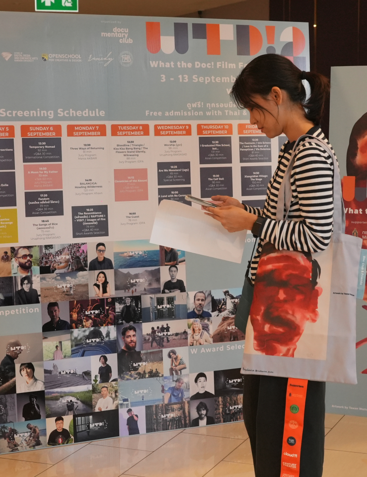 A woman in a striped shirt with a tote bag over her shoulder uses her smartphone. She stands in front of two large banners, one of which displays a schedule for the 2026 edition of What the Doc!, and one of which displays a graphic of a man's face (the same graphic which is on her bag) behind the words What the Doc! 2