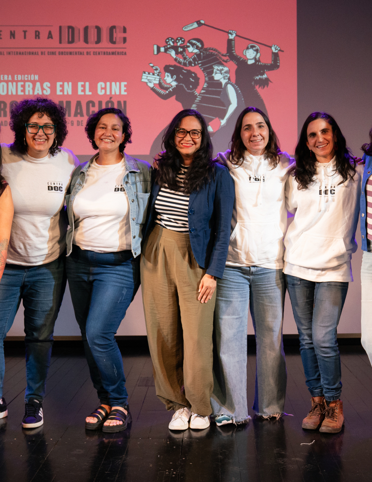 Seven women stand in a row on a black stage, in front of a screen which displays the word "CENTRADOC" and a graphic of women filmmakers over a red background. Four of the seven wear CentraDoc merch, two wear striped tops, and the woman on the left wears an orange, patterned garment