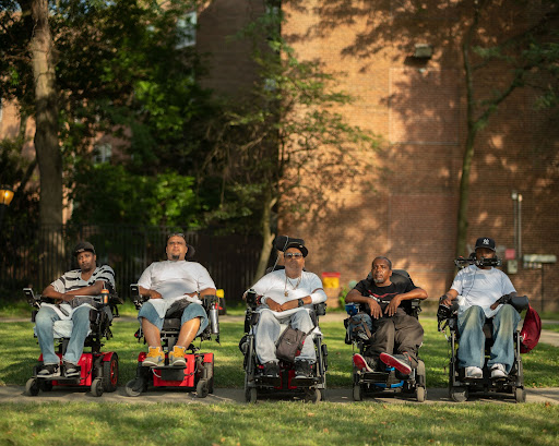 Five Black and brown men (from left to right: Vince, Jay, Tito, Pete and Var) sit in wheelchairs outside, all lined up in a straight horizontal line facing the camera. They wear T-shirts and sneakers and some wear baseball caps. They all look into the camera with serious expressions.