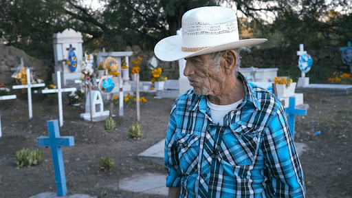 An elderly man in a white sombrero and blue checked shirt stands in a cemetery and directs his gaze downward. Behind him are graves decorated with flowers and balloons.