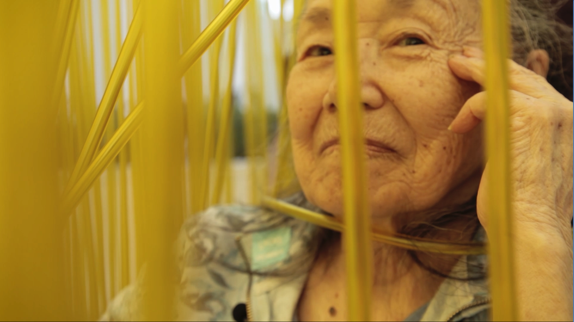 Rose Tajiri, an elderly Asian woman, poses amid an art installation comprised of long yellow strands. From Rea Tajiri's “Wisdom Gone Wild”. Photo courtesy of IDFA